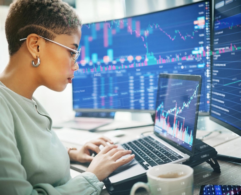 Side View Of Woman Working On Laptop And Two Curved Monitors With Stock And Financial Information On Screen